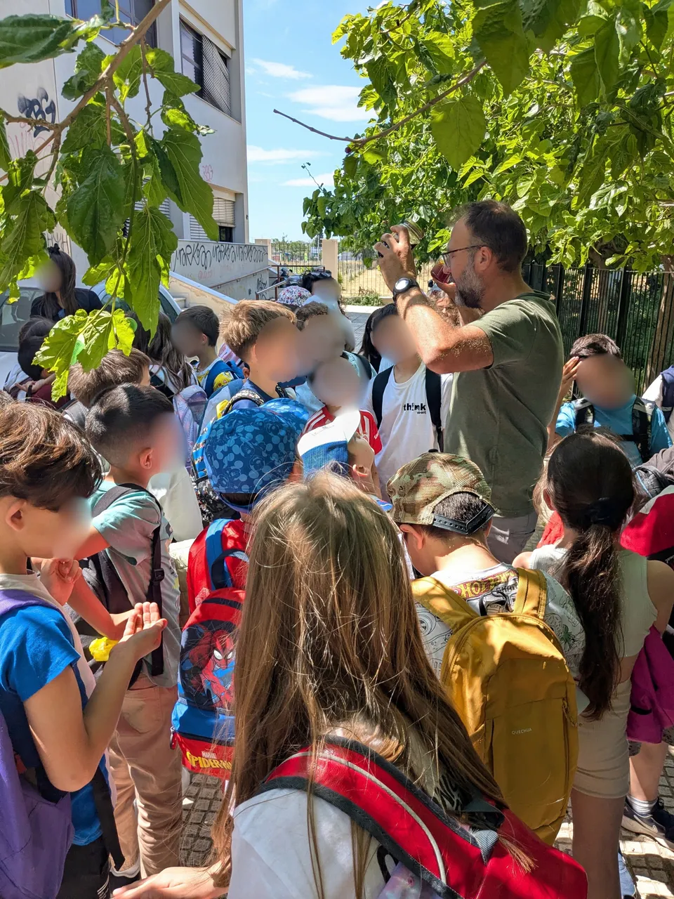 Ομάδα παιδιών παρακολουθεί υπαίθρια παρουσίαση κάτω από δέντρα.
